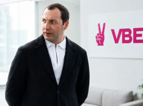 Man in a black blazer stands in a bright, modern office lobby with a pink VBET sign on the wall behind him.