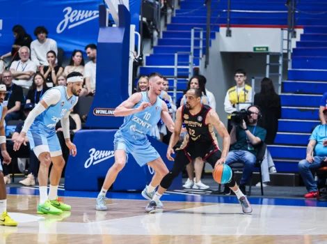 Basketball player in black jersey dribbles toward the basket as blue team players guard him on a glossy court. Audience seated in blue stands behind them.