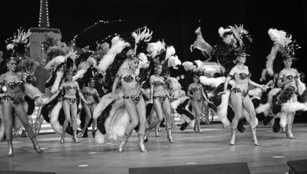 Lido de Paris Group of showgirls in feathered headdresses and glittering bikinis performing a chorus line on a stage.