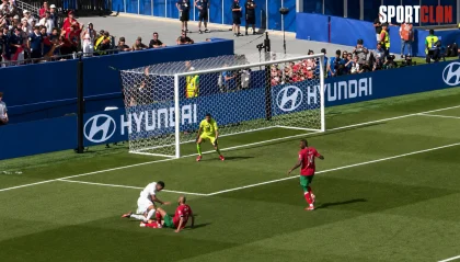 Two players on the ground near the goal with a white player helping a red-and-green teammate as the ball is not visible, a goalkeeper in bright yellow guards the net, and a defender in red and green stands nearby while fans watch from the stands and Hyundai banners line the field.