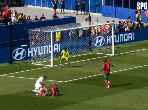 Two players on the ground near the goal with a white player helping a red-and-green teammate as the ball is not visible, a goalkeeper in bright yellow guards the net, and a defender in red and green stands nearby while fans watch from the stands and Hyundai banners line the field.