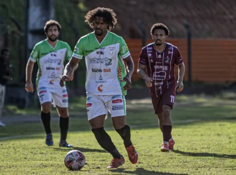 Three soccer players on a grassy field; the central player in green-white controls the ball as two opponents watch nearby.