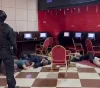A heavily armed officer in black tactical gear with 'OMOH' on the back stands over several people lying face down on a checkered floor in a gaming/computer room as a security operation unfolds.