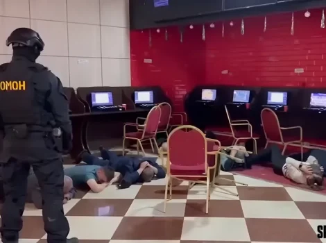 A heavily armed officer in black tactical gear with 'OMOH' on the back stands over several people lying face down on a checkered floor in a gaming/computer room as a security operation unfolds.