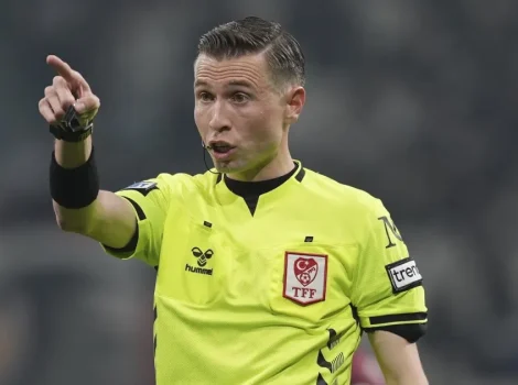 Soccer referee in a bright yellow kit points to indicate a decision during a match, headset mic visible against a blurred stadium background.