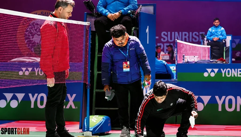 beards Indoor badminton court scene: a man in red near the net, a kneeling opponent in black, and an official in blue with a lanyard nearby; Yonex branding visible.
