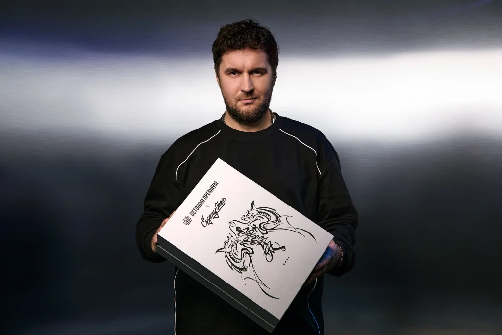 фото Portrait of a man with dark hair and beard in a black sweatshirt, holding a white illustrated booklet with a dragon drawing against a blue-gray gradient background.