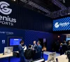 Genius Sports booth at a trade show with a large logo and a blue wall reading 'Discover more' and several attendees talking at a wooden desk setup.