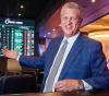 Smiling man in a blue suit sits at a casino bar with sports betting boards in the background.