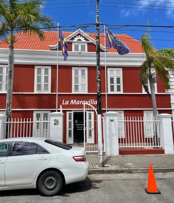 фото 216 Red two-story building with white shutters labeled 'La Maravilla', flanked by palm trees, a white fence, and purple flags on a sunny street with a parked white car nearby.