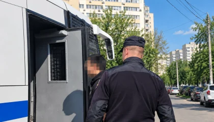 Police officer guiding a detainee into a barred police transport van on a city street; detainee's face blurred.