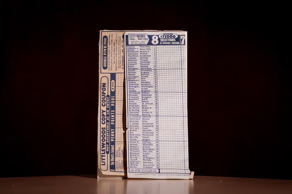 Vintage Littlwoods football pools coupon form with a grid of team fixtures, standing upright on a dark surface against a black background