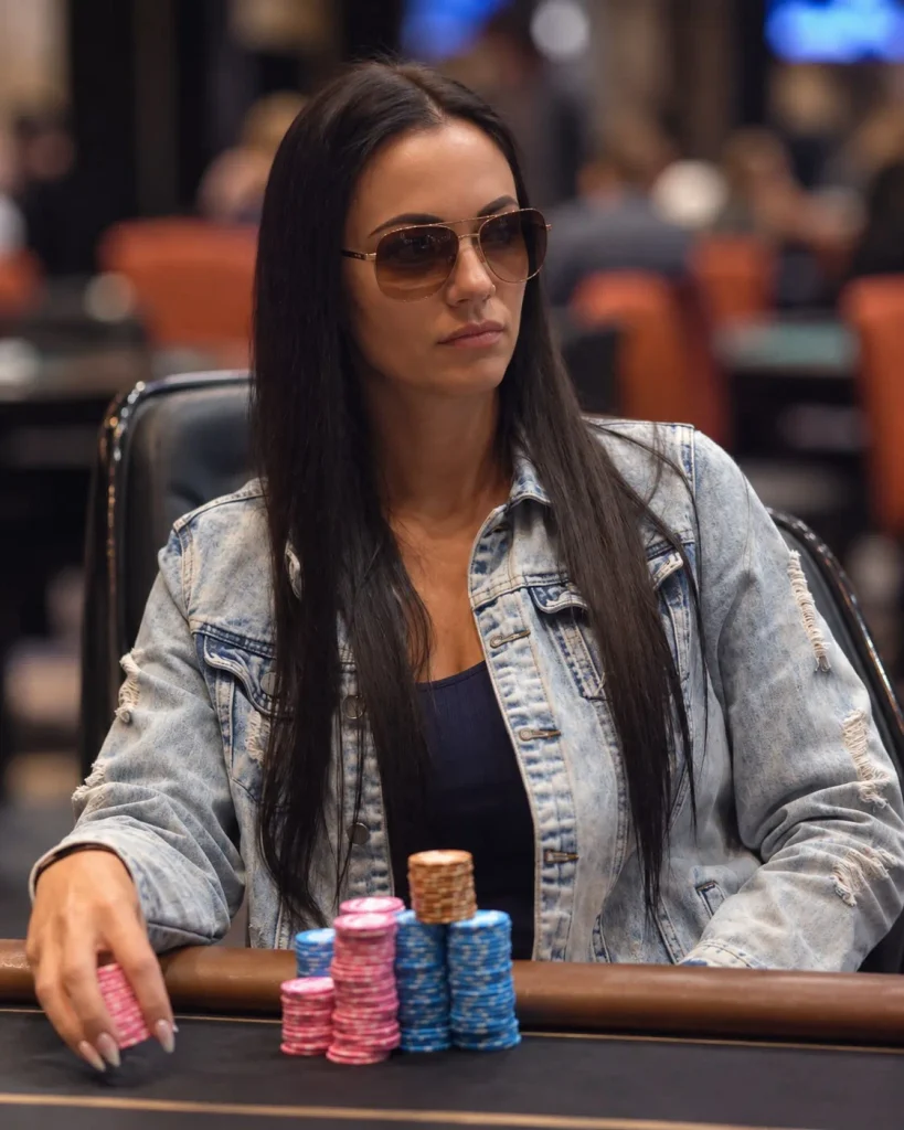 Джейми Кемпф Woman in sunglasses and a denim jacket sits at a poker table with stacked pink, blue, and brown chips in front of her.